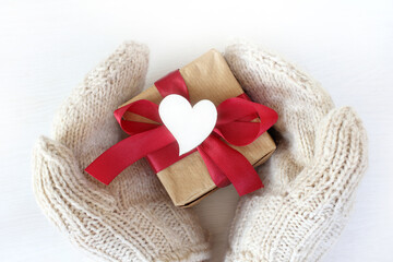 hands in white knitted mittens hold a gift with a red bow decorated with a heart symbol, top view. Congratulations on the Day of lovers