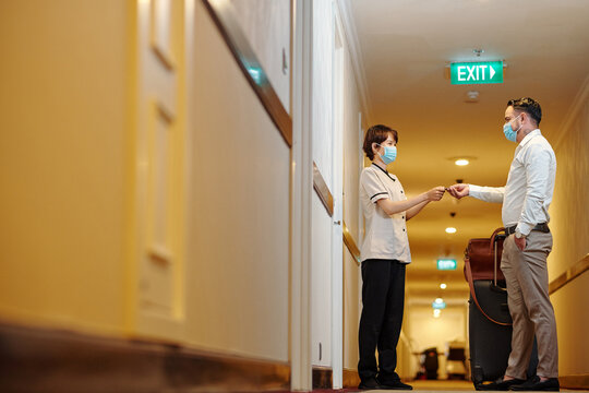 Young Female Hotel Worker In Medical Mask Giving Electronic Key To Guest With Big Suitcase