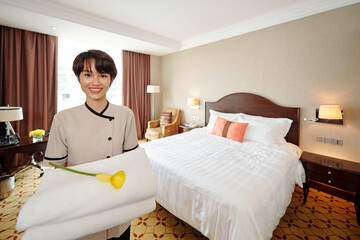 Portrait of pretty smiling young Vietnamese maid standing in hotel room with stack of towel and beautiful calla lily flower