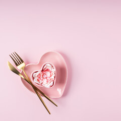 Romantic table setting for Valentines day. Love layout made with golden spoon and fork, heart shaped plate and rose petals on pink background. Minimal Valentines day or wedding dinner. Flat lay.