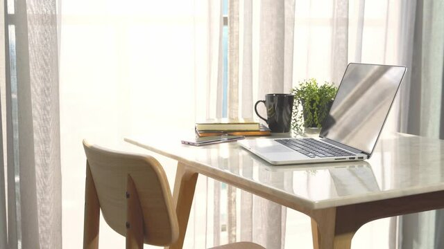 Beautiful workspace - online remote work from home concept. A wooden table with computer laptop, smartphone, coffee cup beside the window balcony with sun light shine through the curtain.