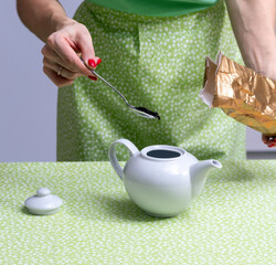 A red-lacquered hand holds a spoon of tea leaves and drops them into a white teapot from the bag.