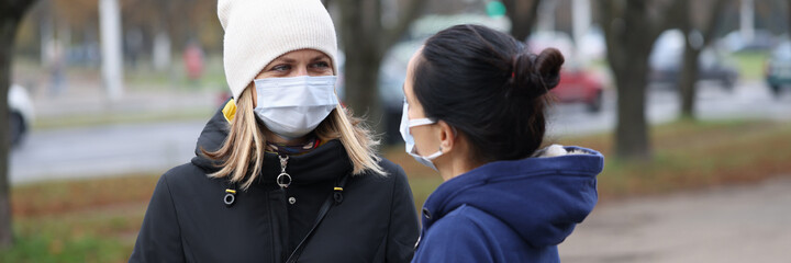 Fototapeta premium Two young women communicating in protective face masks outside. Safety rules during the covid-19 pandemic concept