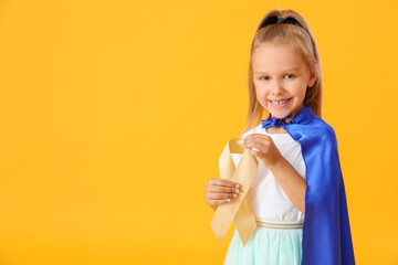 Little girl wearing superhero costume and with golden ribbon on color background. Childhood cancer...
