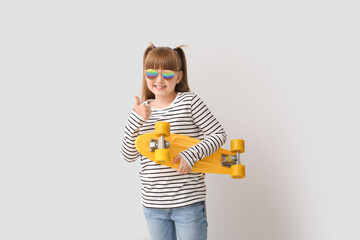 Cute little girl with stylish sunglasses and skateboard showing thumb-up gesture on light background