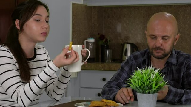 A loving young couple together drink tea or coffee with cookies sitting in the kitchen. The woman is telling a story, the man is chatting on the phone.
