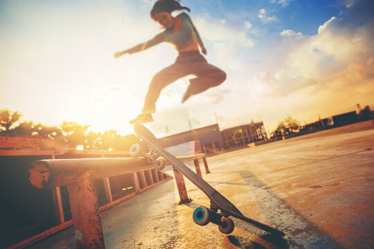 Young Woman Practising Skateboarding At Skate Park. Women Try To Playing Skateboard Jumping At The Roll Bar With Sunset Background.