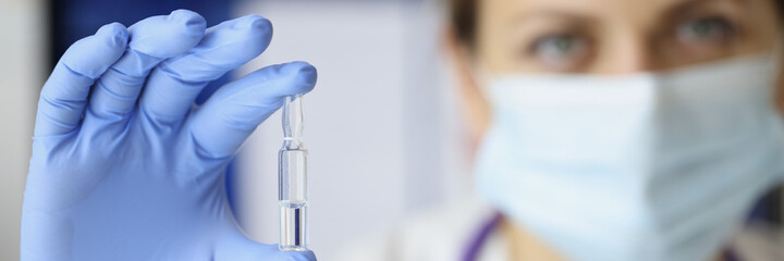 Doctor in rubber gloves and medical mask on face holding ampoule of medicine closeup. Injection drug administration concept