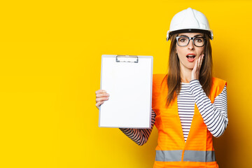 Young woman with a surprised face in a vest and hard hat holds a clipboard on a yellow background....