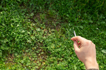 A man's hand holds a cigarette against a background of bright green grass. The concept of the harm of smoking.