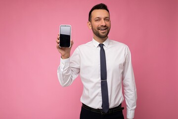 Photo of handsome good looking man wearing casual white shirt and tie isolated on pink background with empty space holding in hand and showing mobile phone with empty screen for mockup looking at