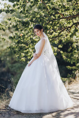 Wedding portrait of a beautiful brunette bride with curly hair in a white dress in nature, against the background of a tree in greenery.