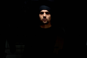 teenager with goatee, in black background, dressed in a sweater and a black hat.Man with...
