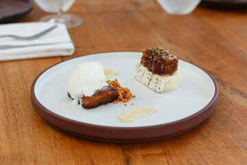 Gourmet dessert, including a honey cake, goat cheese and a honeycomb, served on a flat round stoneware plate with a tall rim. Background of a wood table, set for dinner.