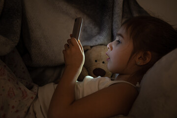 A cute little girl enthusiastically plays on the phone at night in bed instead of sleeping.