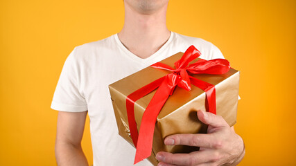 Cropped view of young handsome man holding gift box against colored background. Big gold box with red ribbon. Holidays concept