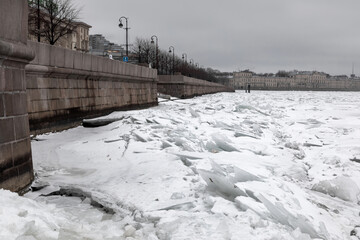 Snowy frozen river with a beautiful old stone embankment. Chunks of ice on the water. Cloudy winter day.
