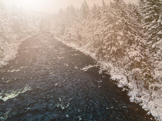 River running through a snow covered wild forest in the freezing winter at sunrise.