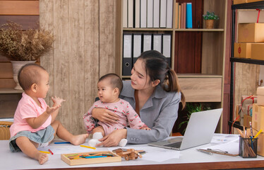 Beautiful Asian single mother happy playing with adorable kids while working at home, children play peekaboo with fun on mother working desk, multi-task young freelancer woman with daughters concept