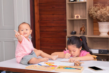 Adorable 2 kids sitting on desk full of crayon colors and white papers enjoy playing together at home, 2 sibling happy drawing with their imagination, elder sister play with naughty younger sister