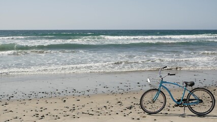Obraz premium Blue bicycle, cruiser bike by ocean beach pacific coast, Oceanside California USA. Summertime vacations, sea shore. Vintage cycle on sand. Clear sky and water waves. Waterfront summer near Los Angeles