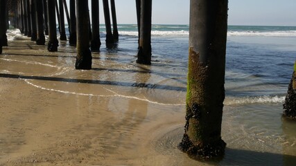 Wooden piles under boardwalk, old pier in Oceanside, California coast USA. Pilings, pylons or pillars below retro vintage bridge, waterfront promenade. Ocean waves, sea water tide and sand beach.