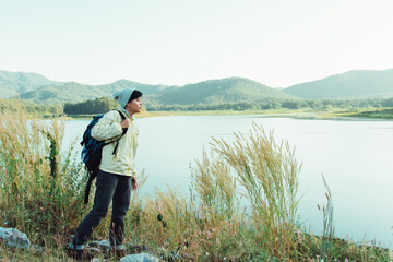 Asian man backpacking in Sweater and grey hat standing on the floor looking Landscape view and mountains at Maetip Reservoir Lamphun Province, Northern Thailand Province in the morning.