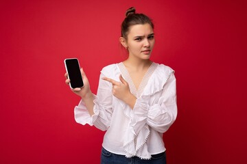 Naklejka premium Photo of beautiful upset young woman good looking wearing casual stylish outfit standing isolated on background with copy space holding smartphone showing phone in hand with empty screen display for