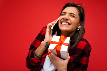 Photo shot of pretty positive young brunette woman isolated over red background wall wearing white casual t-shirt and red and black shirt holding white gift box with red ribbon and talking on mobile