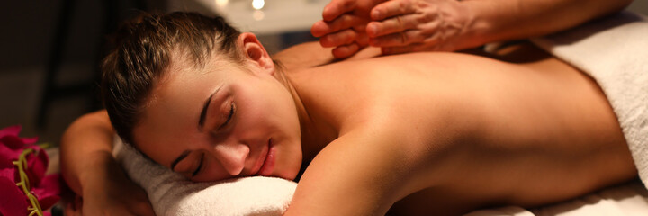 Passive girl lye on massage table covered with white towel and enjoy. Man do back massage with his hand. Candle burn in background.