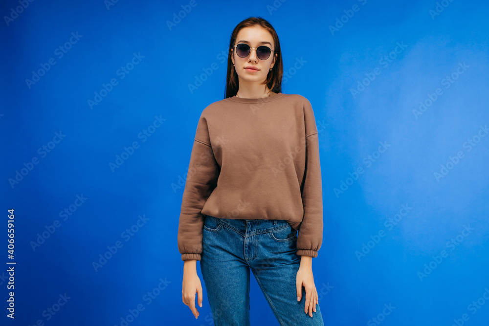 Wall mural Young woman in glasses and hoodie posing in studio on blue background