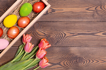 Colorful Easter eggs on a bed of straw in a long wooden box on a wooden table and red tulips. Rustic style, copy space, flat ley