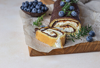 Sponge cake roll with chocolate and cream cheese decorated with chocolate glaze, blueberry and rosemary on parchment, light background. Biscuit swiss roll.