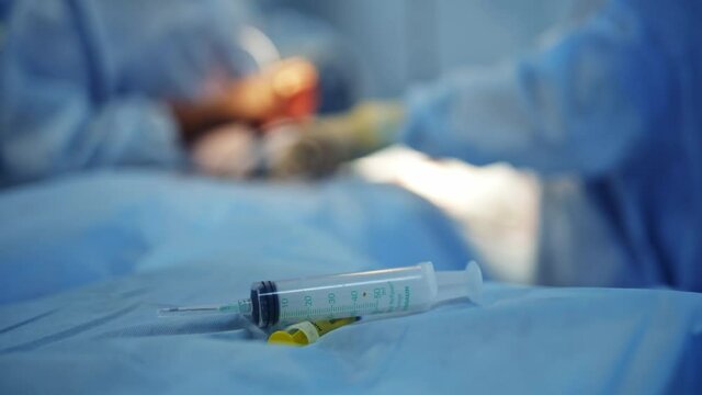 Suringe lays on operation table. Neurosurgery team provides operation on the blurred background. Closeup.
