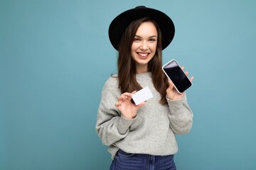 Beautiful smiling happy young brunette woman wearing black hat and grey sweater isolated over blue background holding credit card and mobile phone with empty display for mockup looking at camera