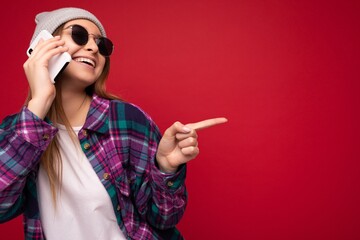photo of beautiful happy positive young blonde woman wearing hipster purple shirt and casual white t-shirt grey hat and sunglasses isolated over red background holding in hand and talking on mobile