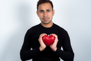 Young man holding a red love heart in his hands in front of his chest, on top of a black pullover, white background. Concept of love, Valentine. 