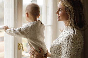 Mother and her little baby son wearing warm sweaters looking at the window