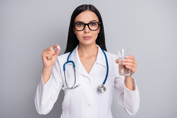 Photo of serious doctor girl hold pill glass water wear specs stethoscope white uniform isolated grey color background