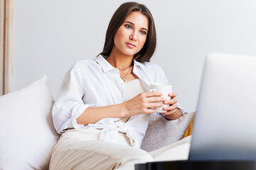 Beautiful brunette in white shirt sits on white sofa, holding a white cup with hot coffee in her hand. A millennial woman looks at laptop screen.