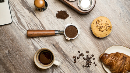 Coffee preparation on the wooden table. Espresso cup with portafilter filled with ground coffee.Muffin and croissant