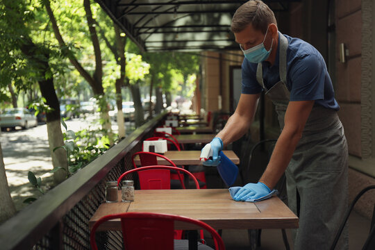 Waiter in mask and gloves disinfecting table at outdoor cafe