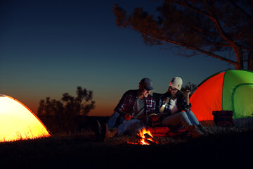Fototapeta na wymiar Couple with flashlight reading book near bonfire at night. Camping season