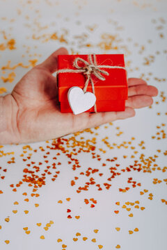 Man heand with small red gift box with golden glitters around. Red present with heart on the rope on white background. Saint Valentine's Day present. Heart-shaped golden glitters.