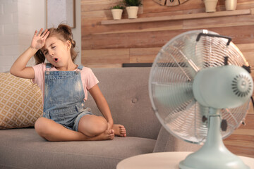 Little girl enjoying air flow from fan on sofa in living room. Summer heat