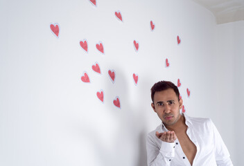 Young man blowing on his hand with many paper hearts flying behind him, white background. Concept of spreading love, Valentine