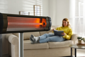Woman reading book in living room, focus on electric infrared heater