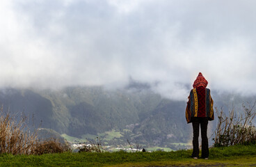 Fototapeta premium Person with hoodie, standing at viewpoint, mountains and clouds, Furnas village, Sao Miguel, Azores islands.