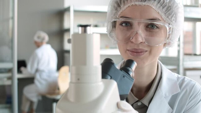 Close up of young Caucasian woman wearing disposable medical hat, goggles and overall doing research using microscope in lab then looking at camera and smiling