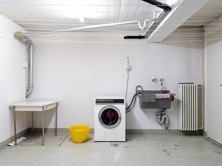 Shared apartment building laundry room with washing machine in used wash sink and piping wide angle...